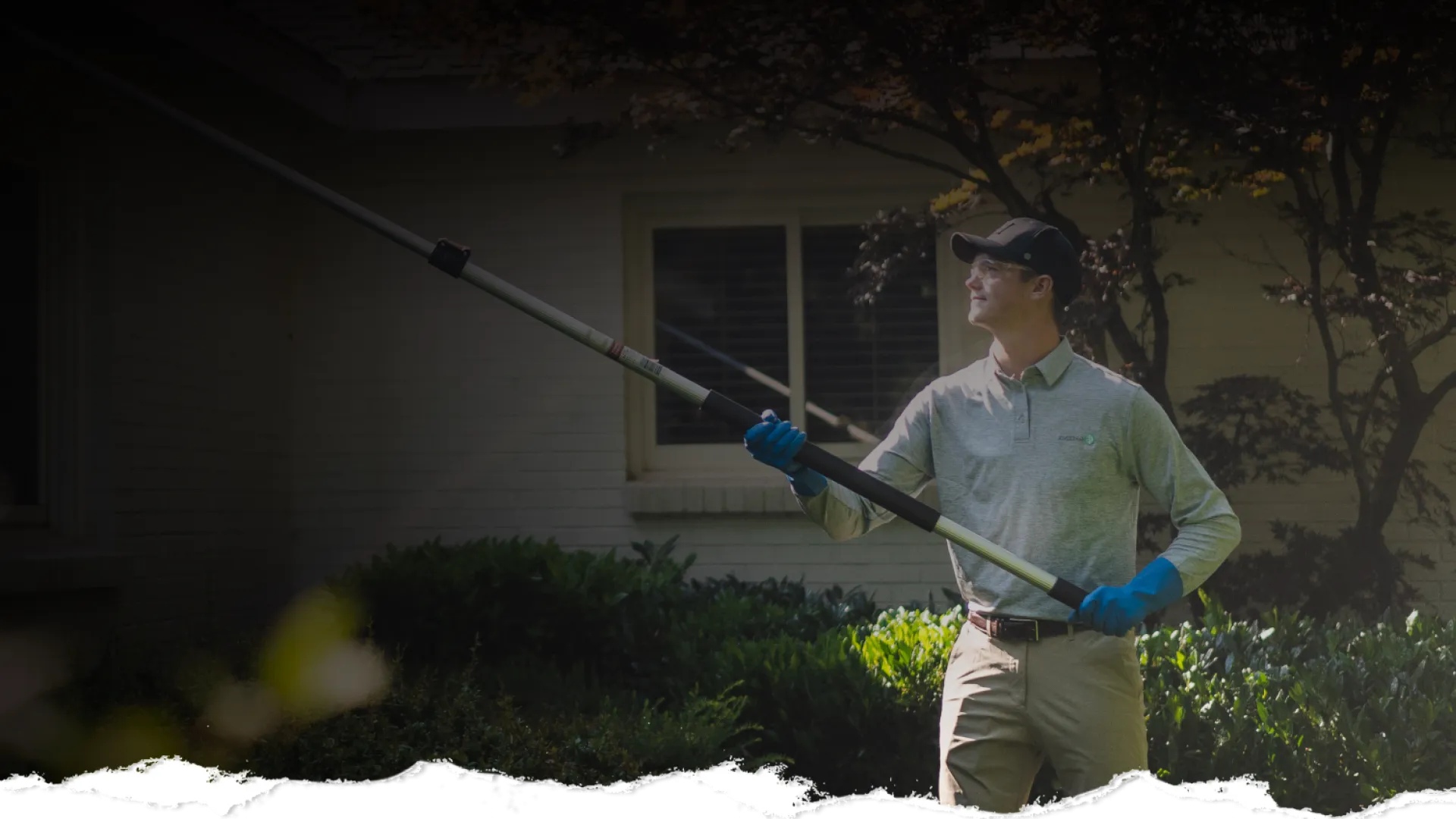 Man in gloves using a long pole tool to trim tree branches outside a house on a sunny day