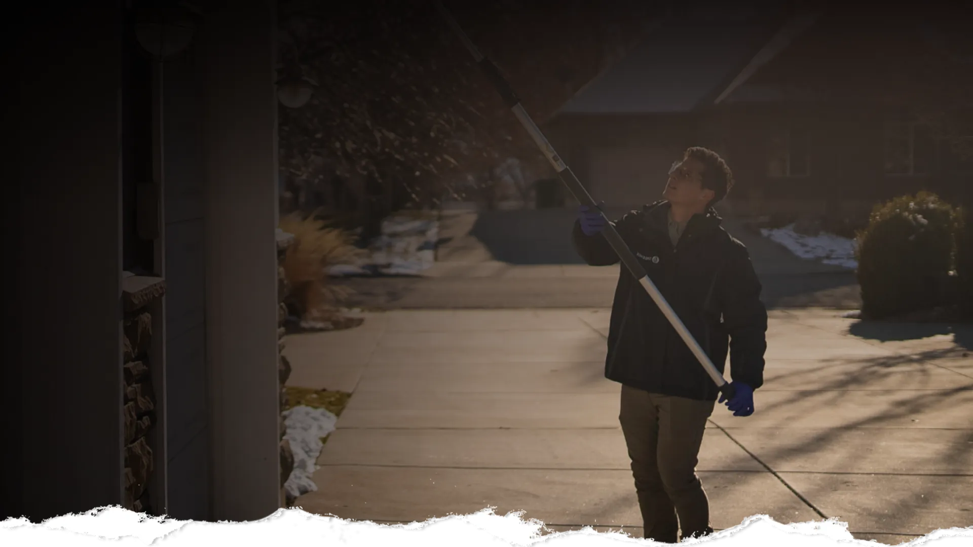 Person wearing gloves uses long pole tool to clean gutters outside a house on a sunny winter day.