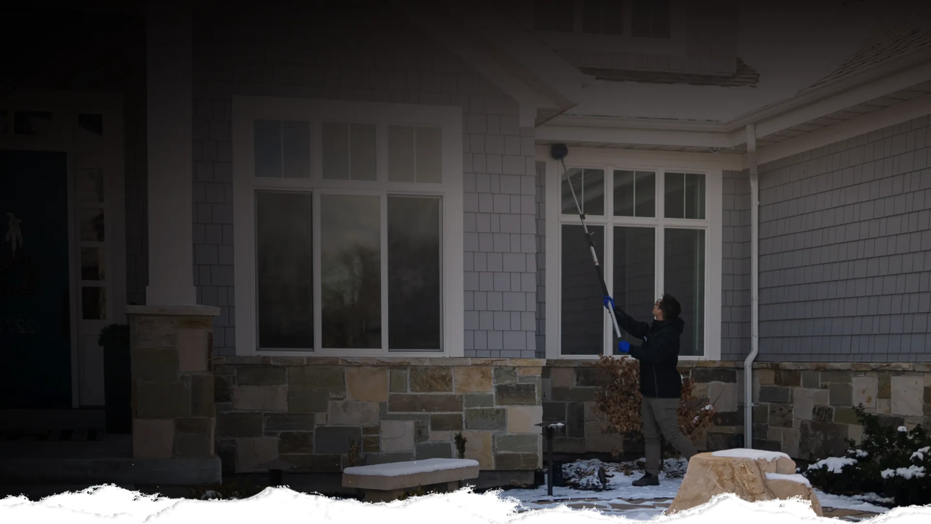 Person using a long-handled brush to clean windows of a stone and shingle exterior house in winter snow.