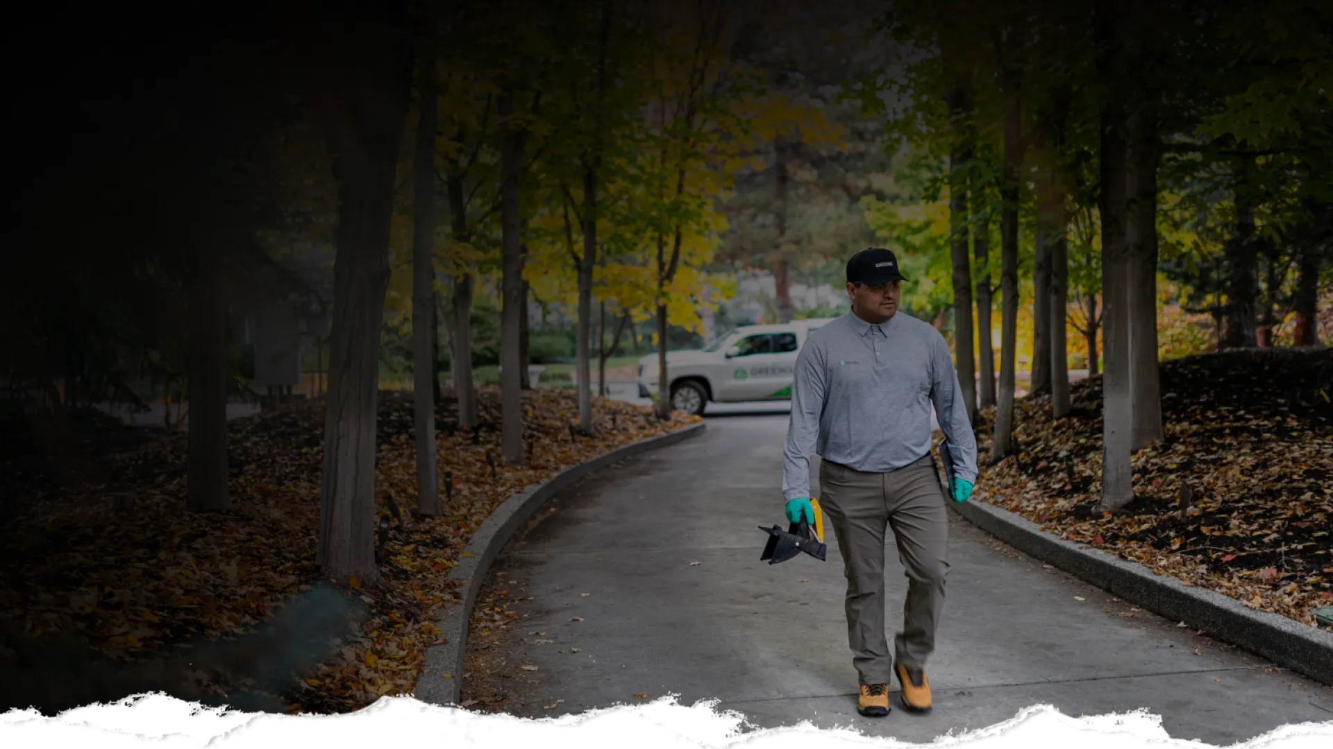 Landscaper walking on a tree-lined pathway with tools and a company truck in the background during autumn.