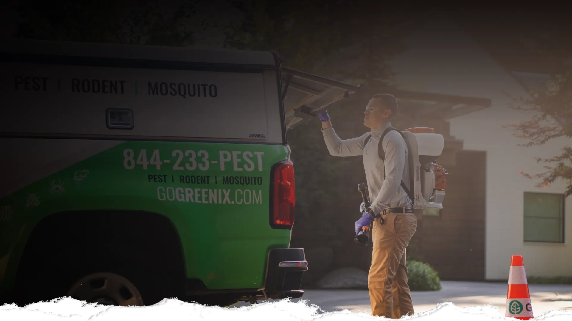 Pest control worker preparing equipment from green service truck next to traffic cone outside a modern home.