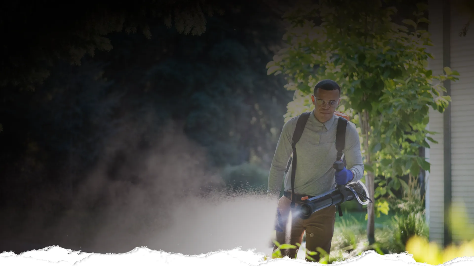 Man wearing protective gear using a fogger to spray pesticide outdoors near trees and a building.