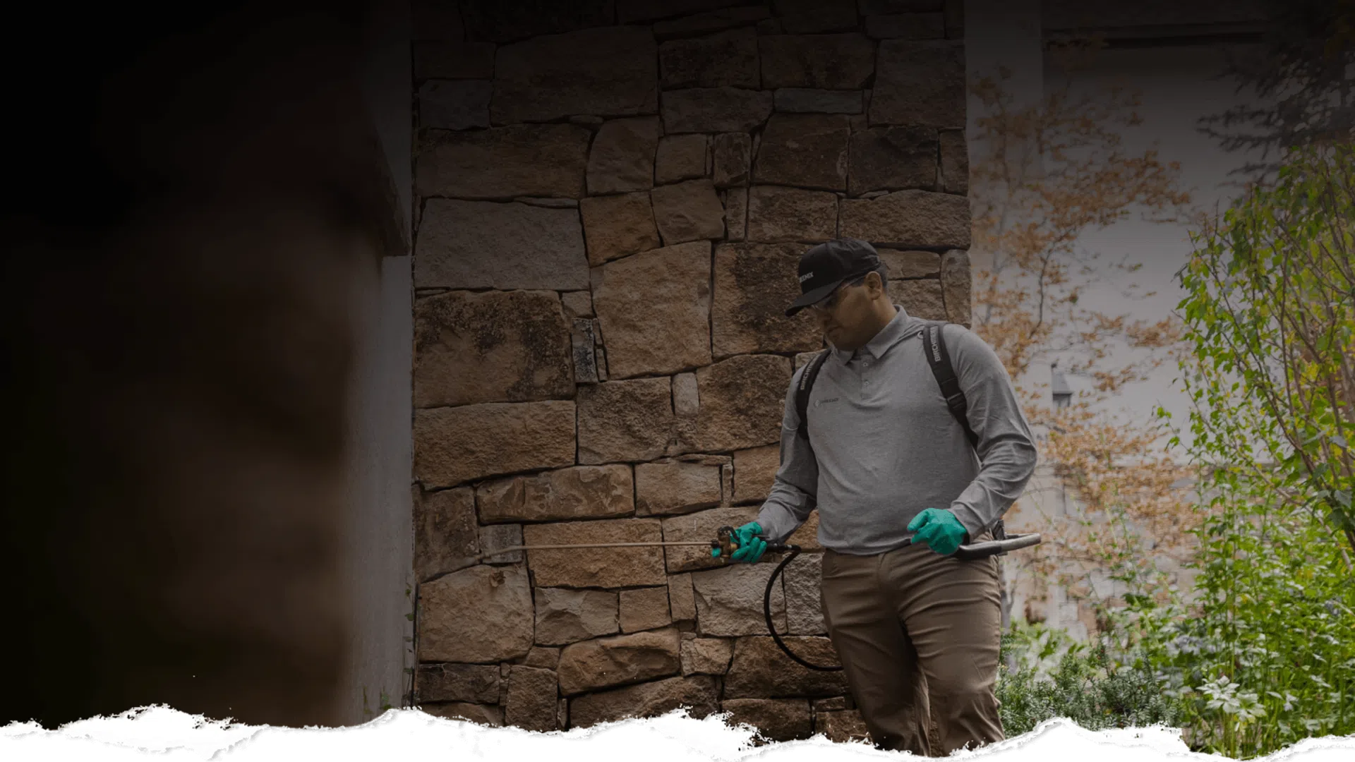 Pest control worker wearing gloves and backpack sprayer treating outdoor area near stone wall.