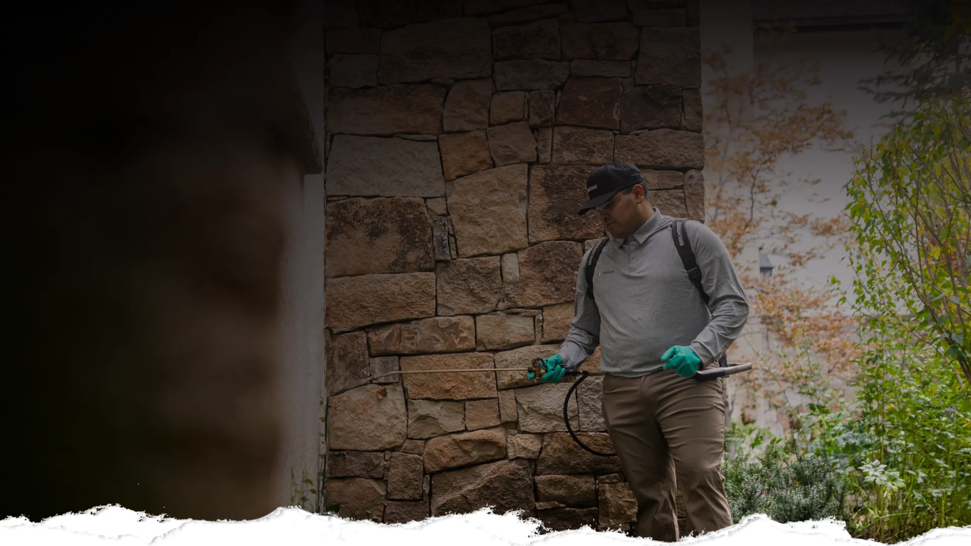 Pest control worker spraying insecticide on a stone wall outside a building with greenery around.