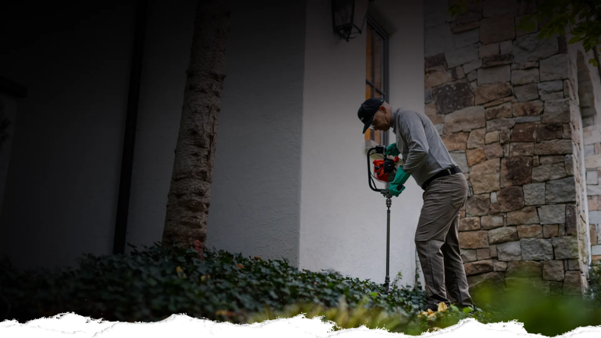 Man wearing gloves and cap using a soil auger drill for gardening next to a stone wall and white house.