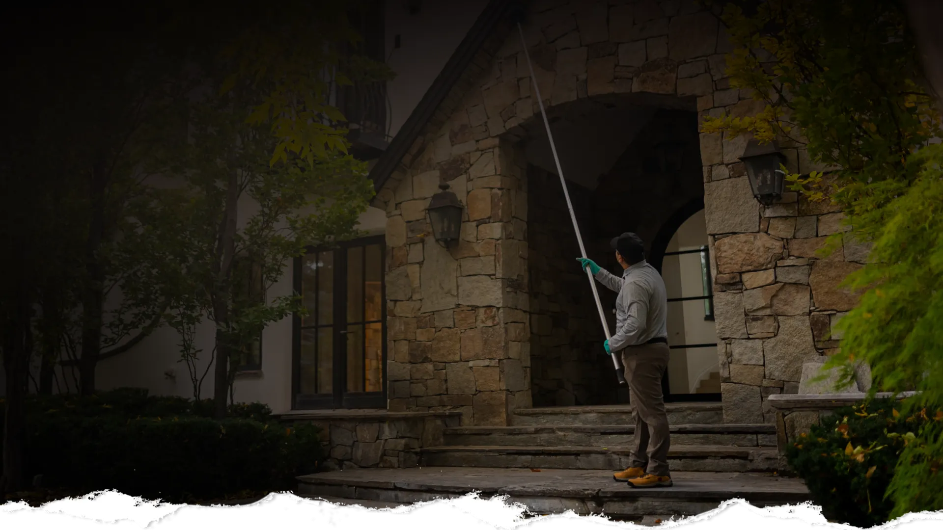 Worker using a long pole to clean or inspect the stone facade of a house entrance on a cloudy day.