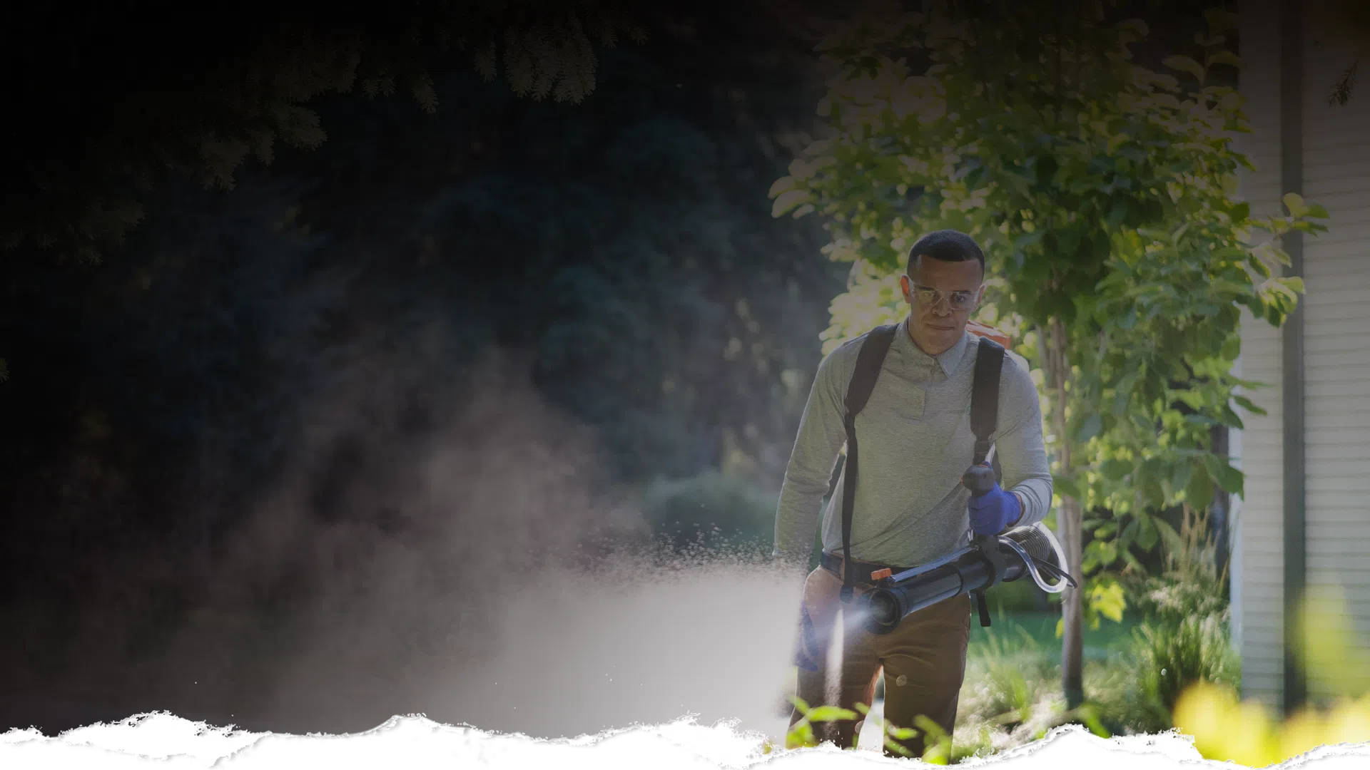 Man wearing backpack sprayer treating plants near a home’s stone and stucco exterior at dusk.