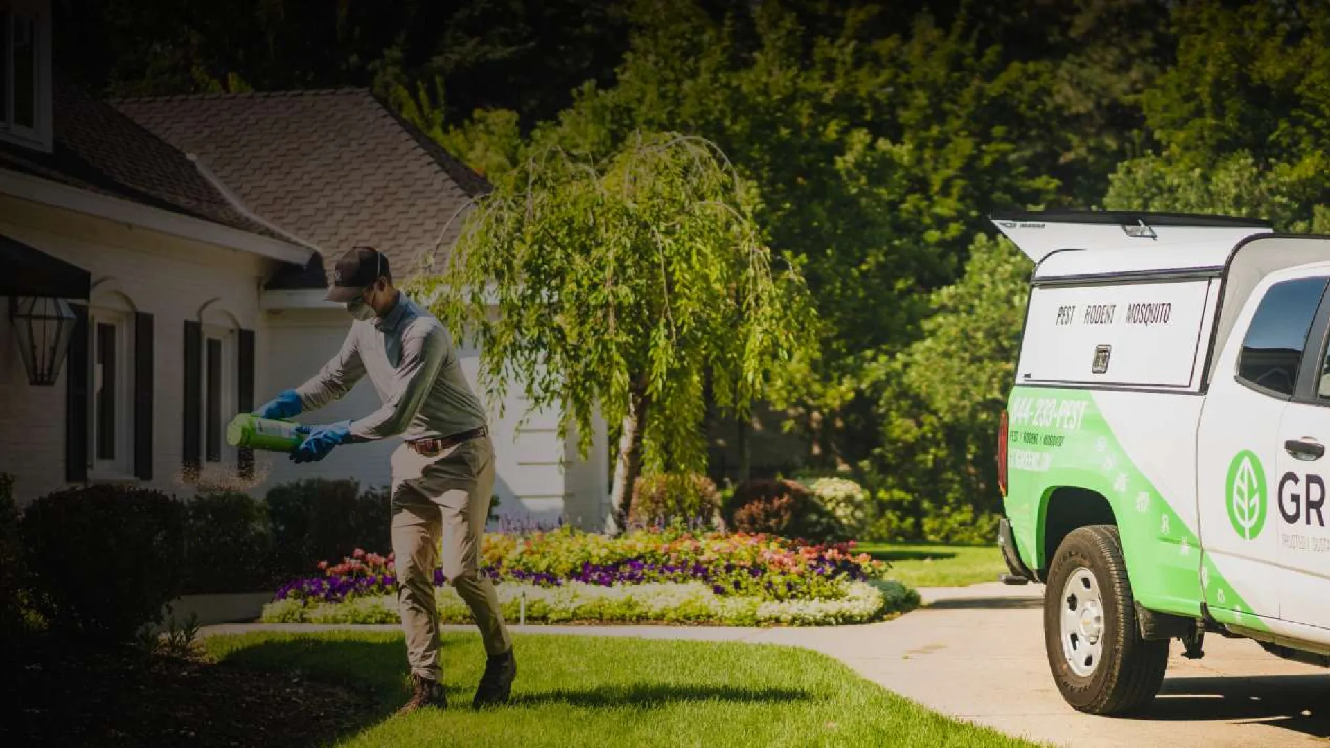 Gardener spraying plants in a residential yard with a Greenix branded white and green service truck parked nearby.
