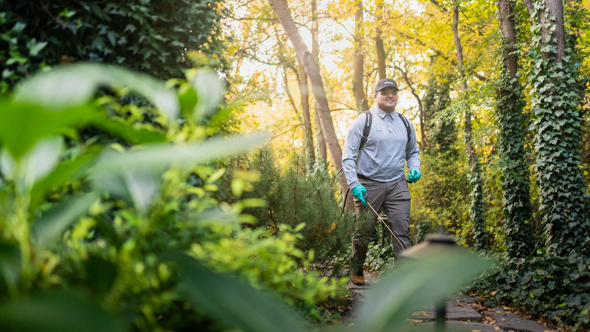 Gardener with gloves and backpack sprayer walking on a sunny garden path surrounded by green plants and trees.