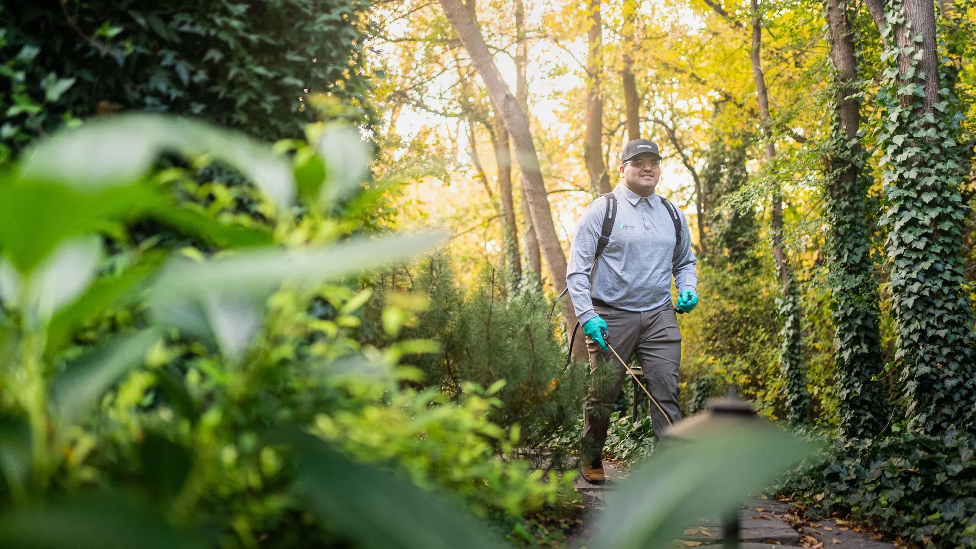 Gardener wearing gloves and backpack sprayer walking through a lush green garden path during daytime.
