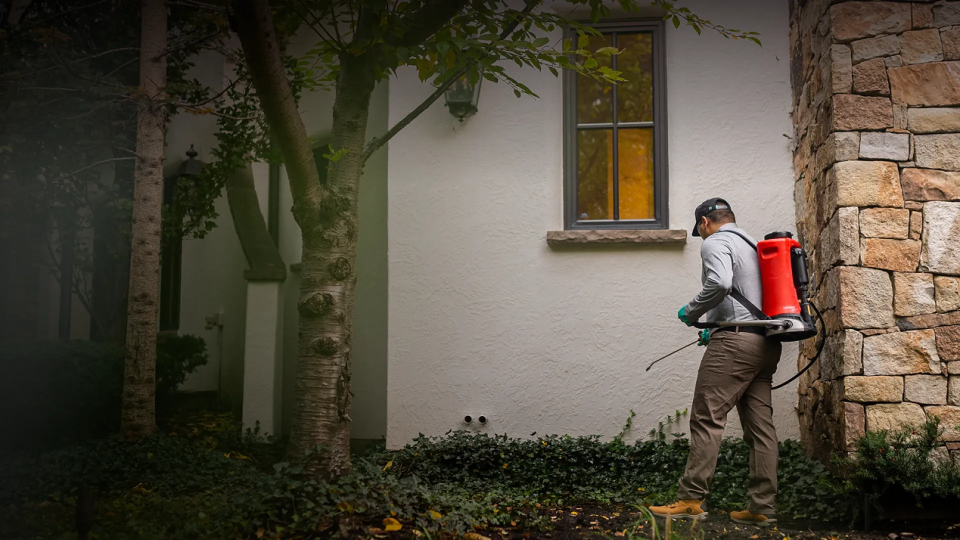 Man wearing backpack sprayer treating plants near a home’s stone and stucco exterior at dusk.
