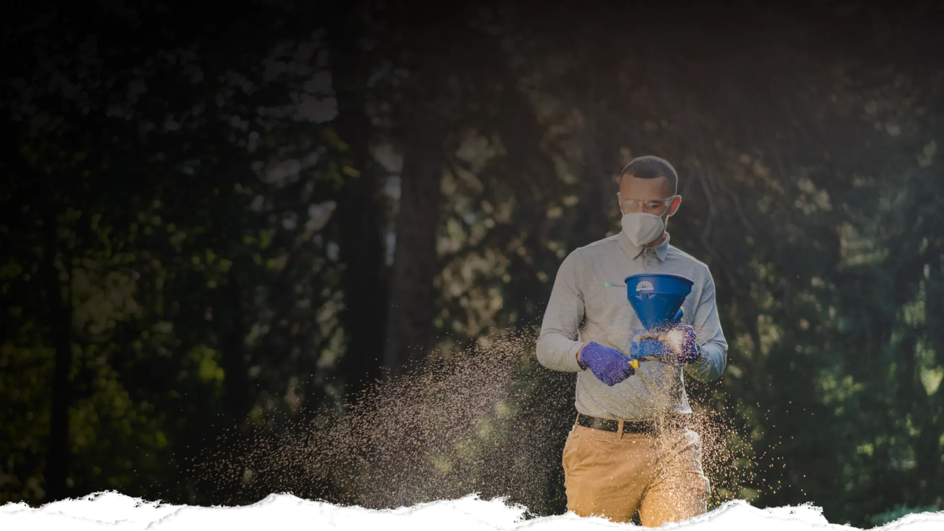 Man wearing mask and gloves spreading seeds or fertilizer outdoors with dense forest in the background