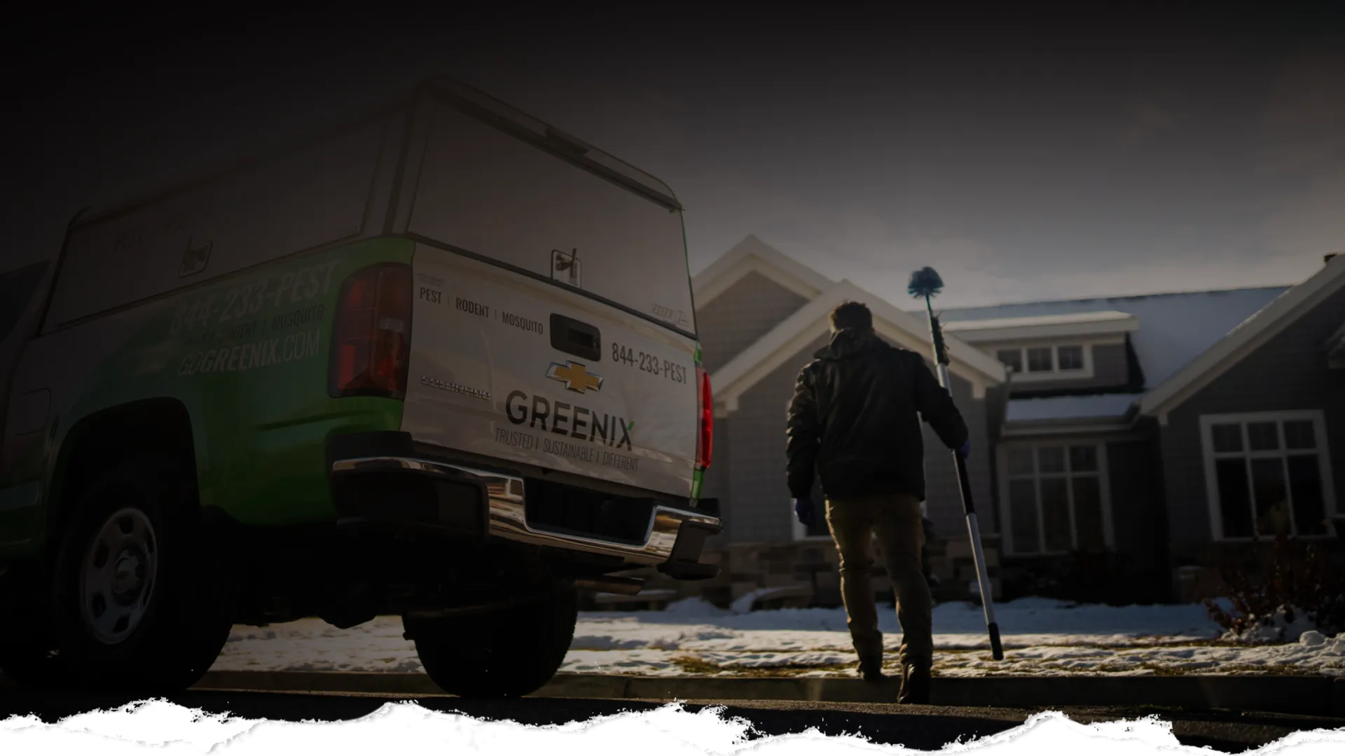 Pest control professional walking with a tool toward a house on a snowy day near a Greenix service truck.