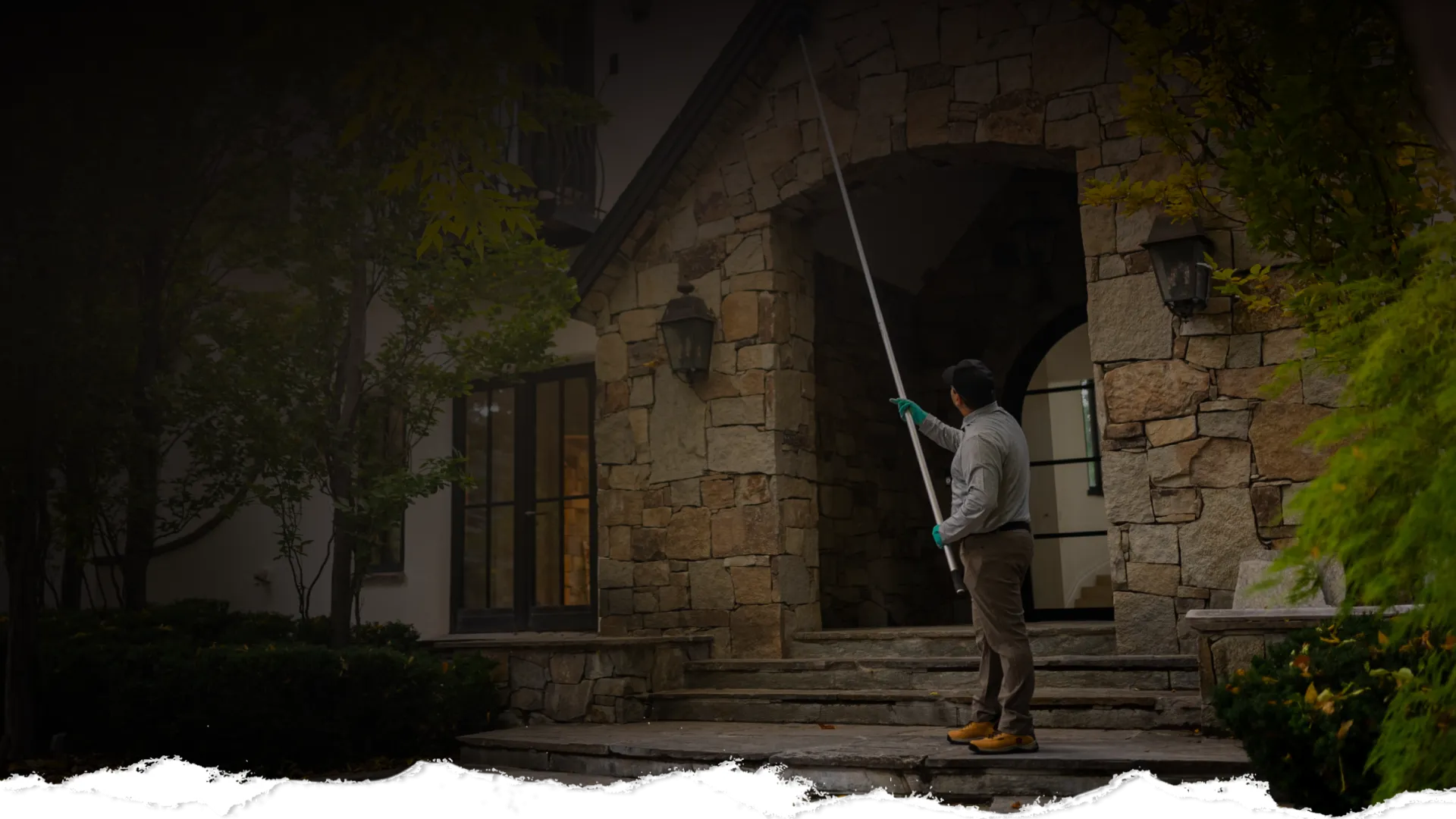Man using a long pole to clean or inspect the stone exterior of a house's entrance at dusk.