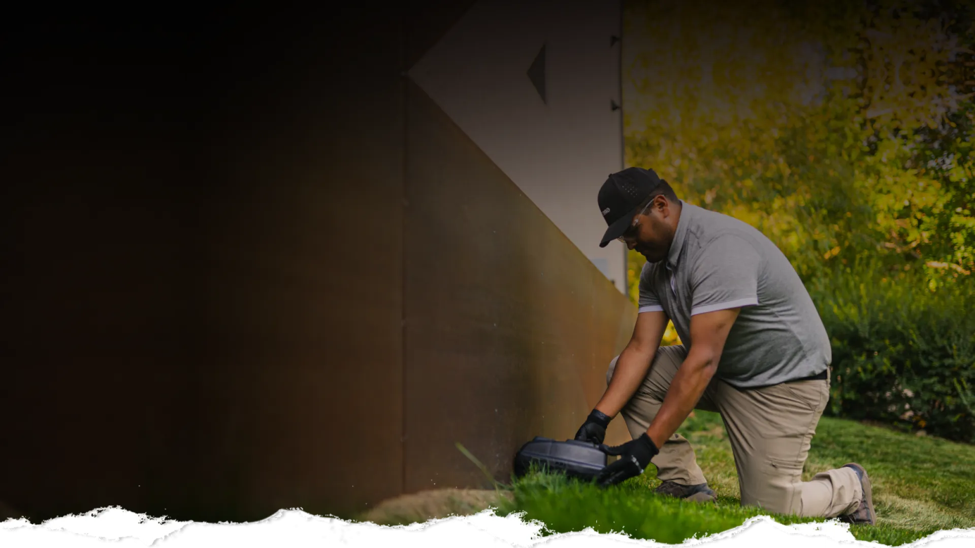 Man in gloves installing or adjusting robot lawn mower outside near a metal wall on green grass.