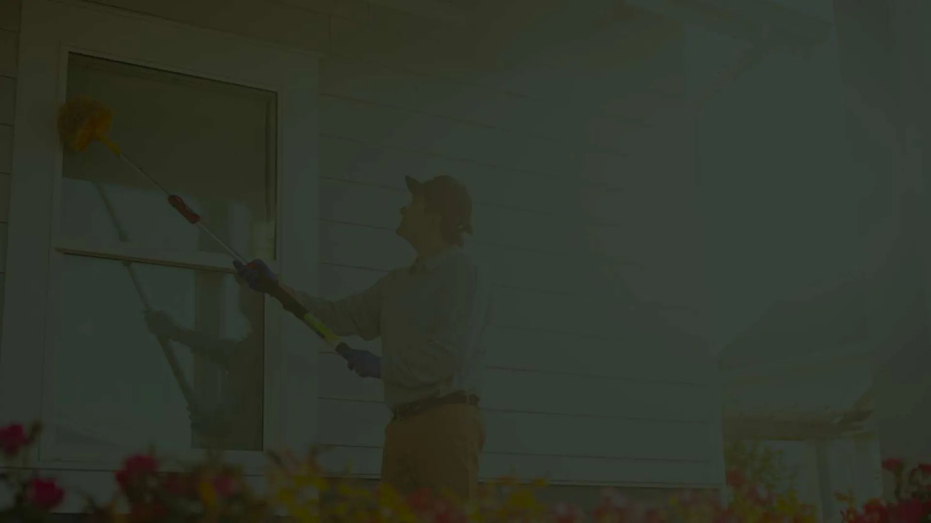 Person cleaning the exterior window of a house using a long-handled brush in daylight.