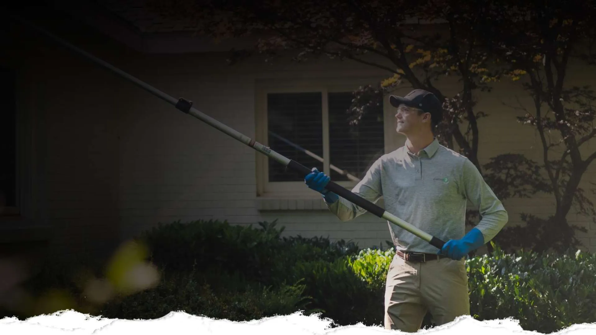 Man wearing gloves and cap using a long pole tool to trim tree branches in a garden