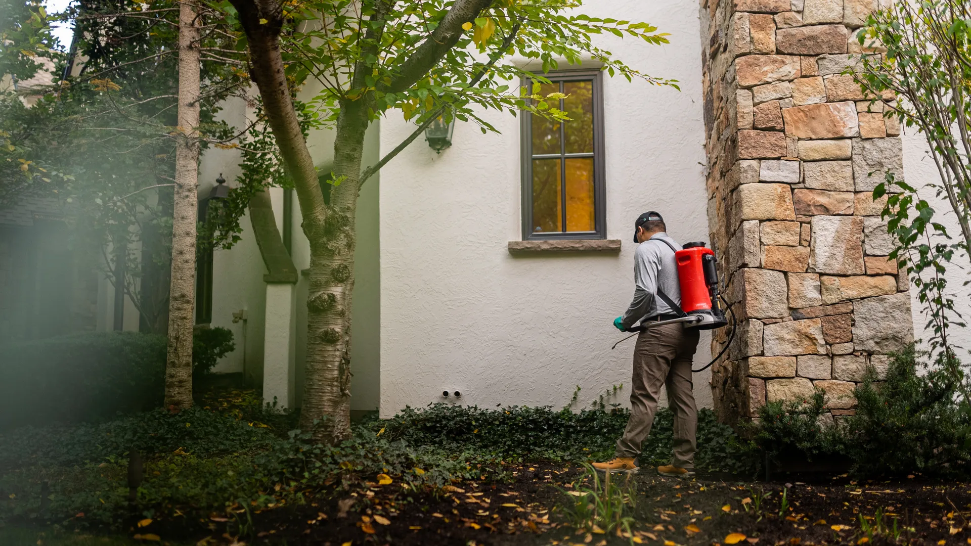Pest control technician spraying pesticide with backpack sprayer near a stone and stucco house in leafy yard.