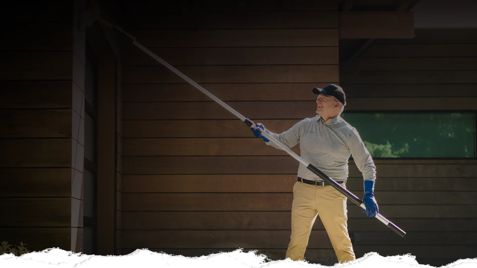 Person using an extendable pole to clean or paint the outside wall of a wooden house wearing gloves and a cap.