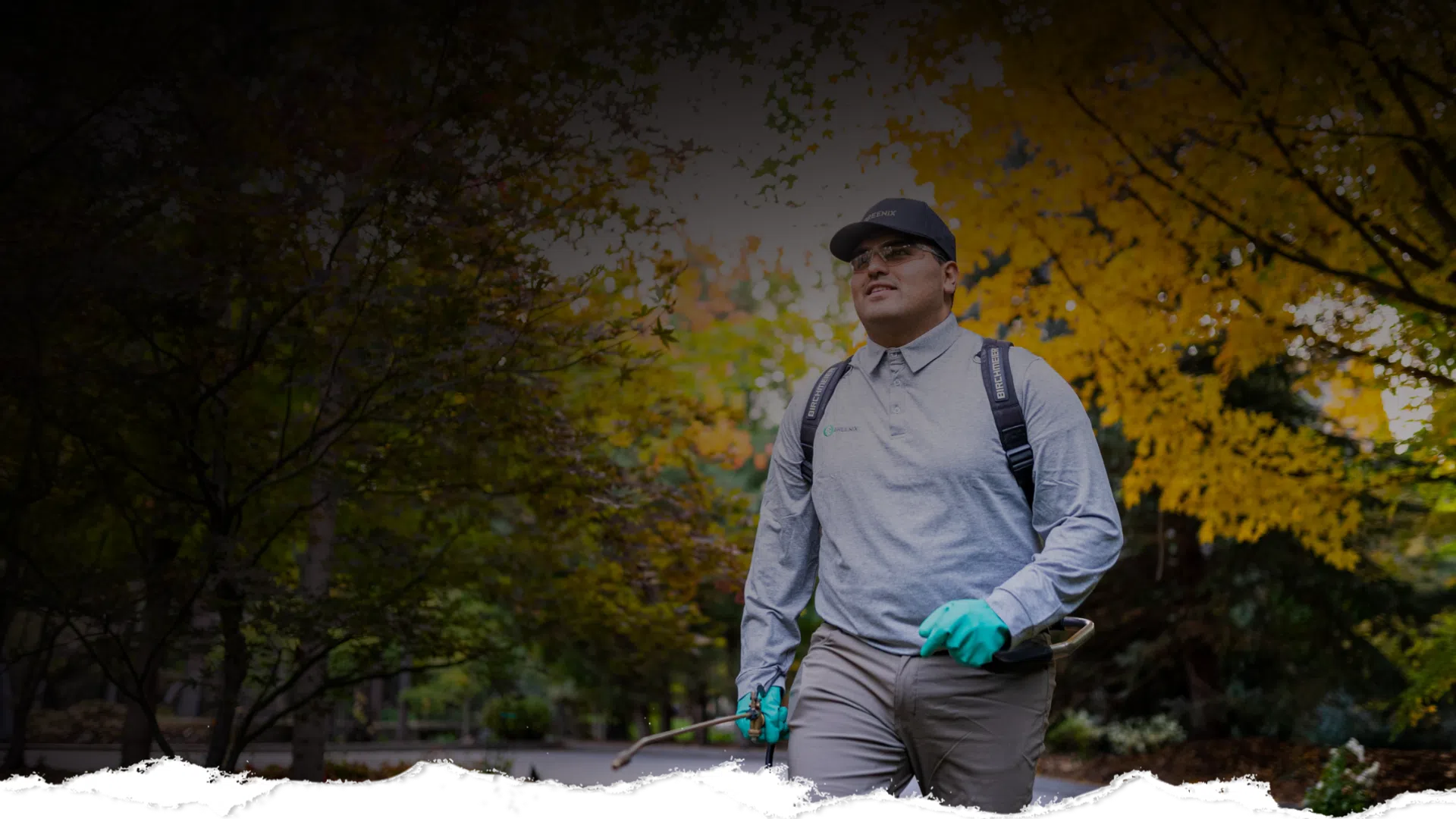 Man wearing gloves and backpack sprayer walking through a park with autumn foliage in the background