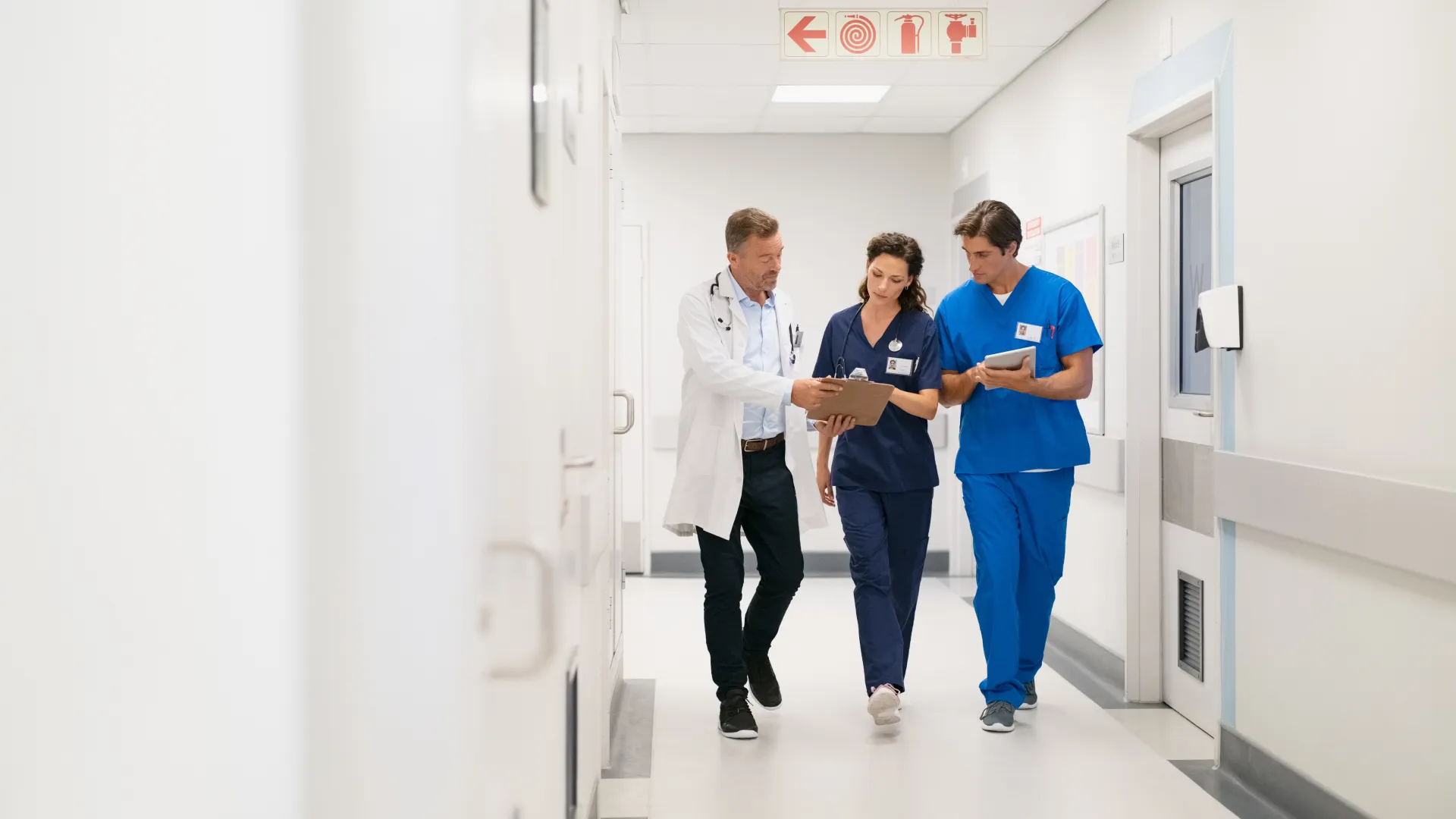 Three healthcare professionals in scrubs and lab coat walking a hospital corridor discussing patient notes.