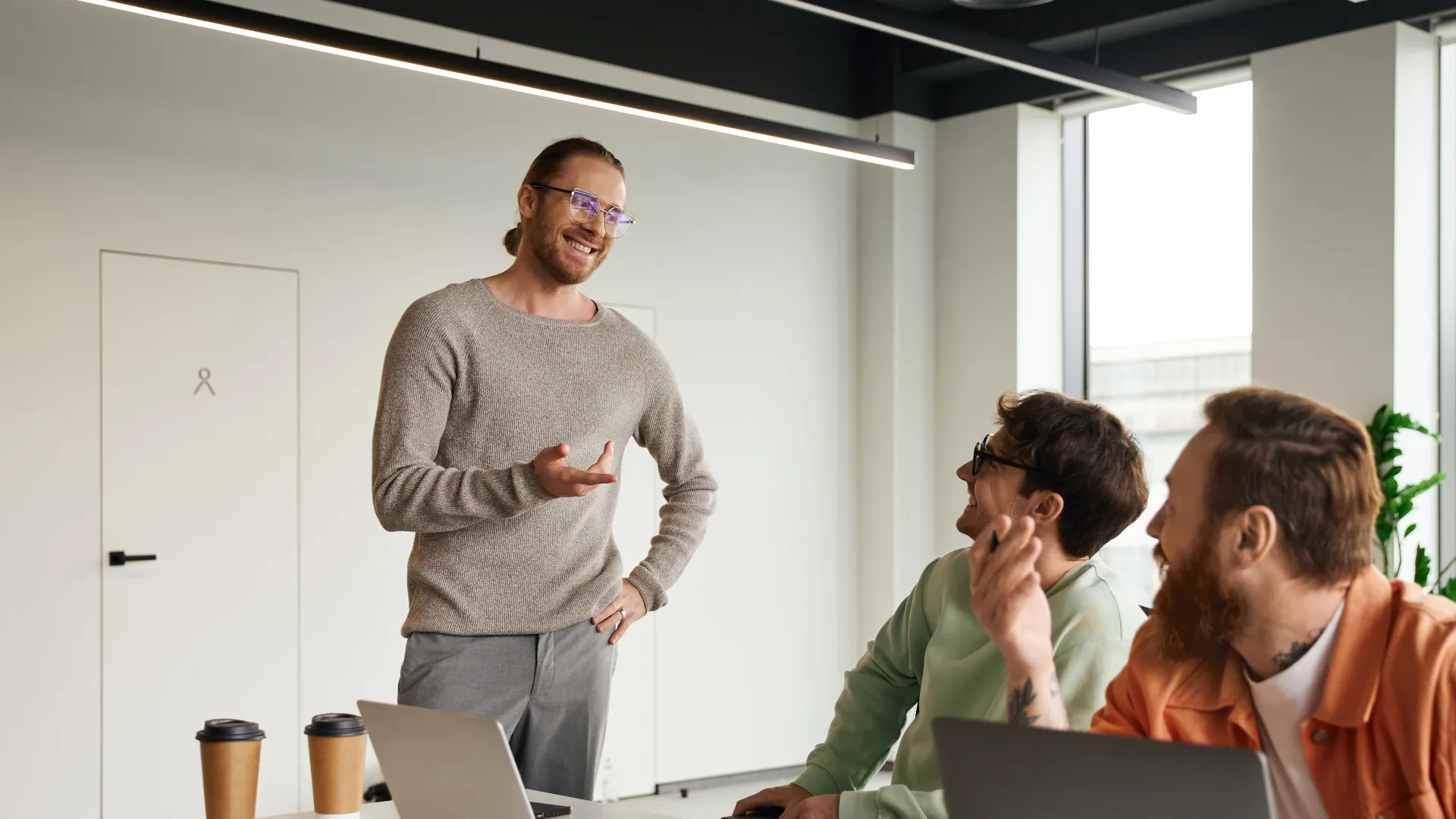 Three colleagues engaged in a casual meeting with laptops and coffee in a modern office space.