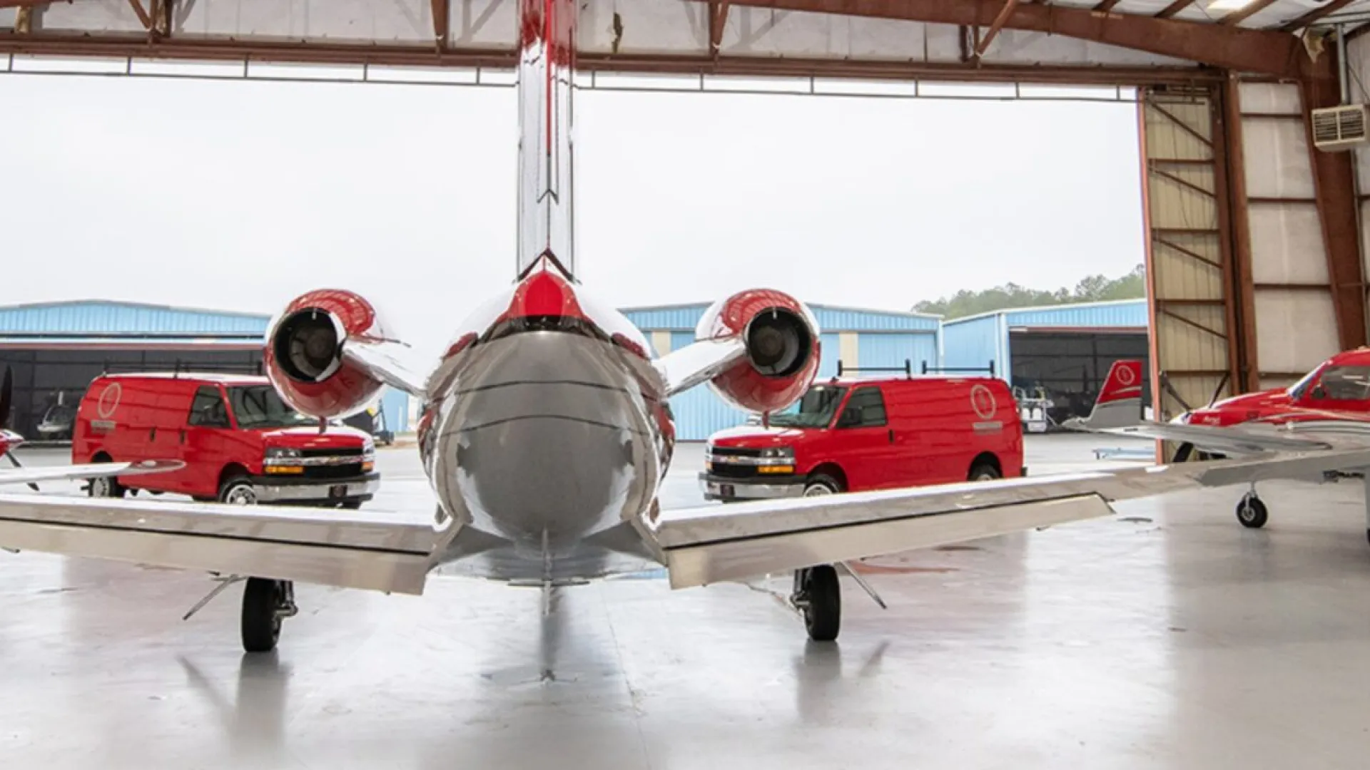 Two red and white small jets parked inside a large hangar with red vehicles in the background.