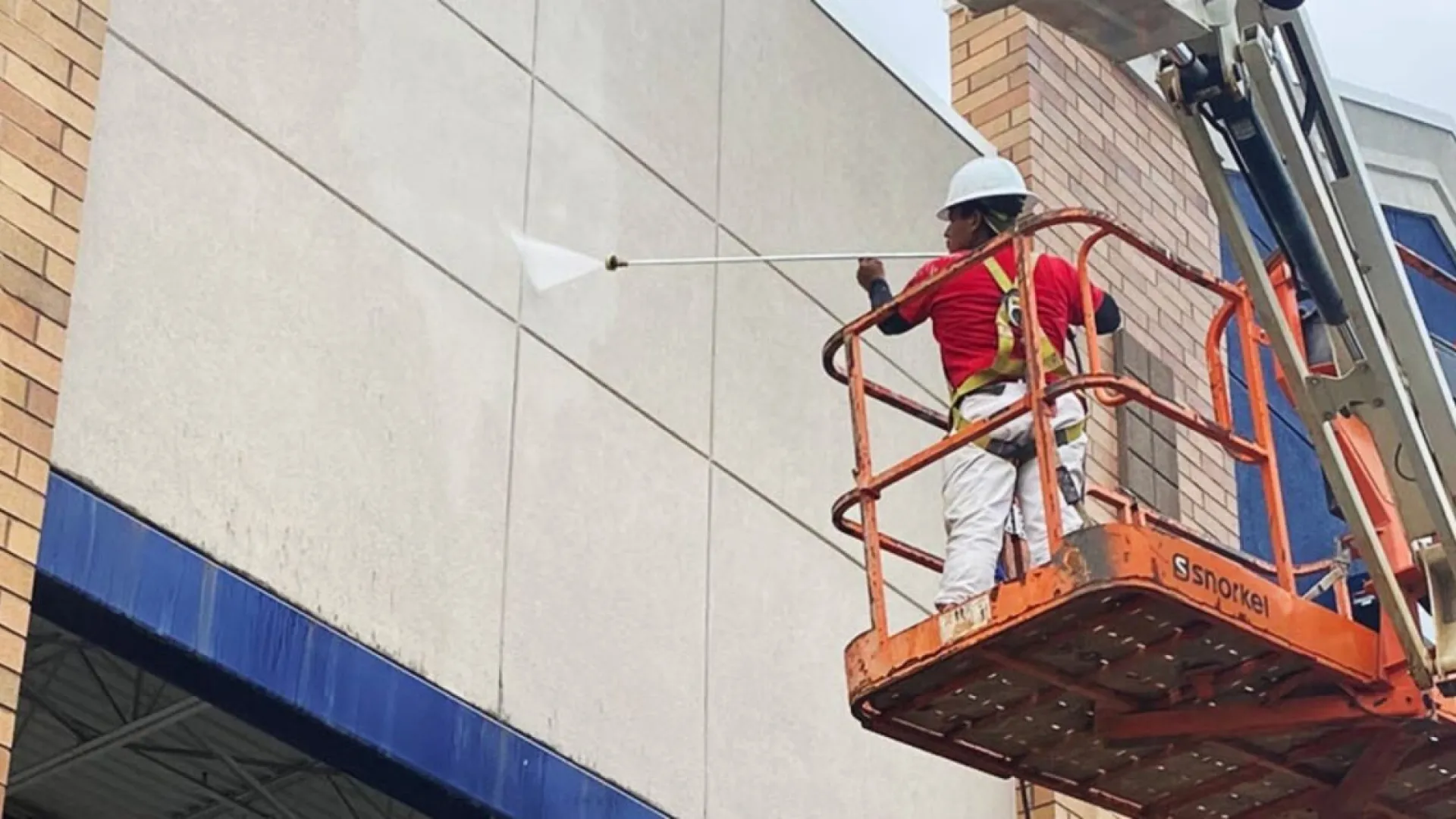 Worker in orange lift cleaning building exterior with high-pressure water spray on a cloudy day.