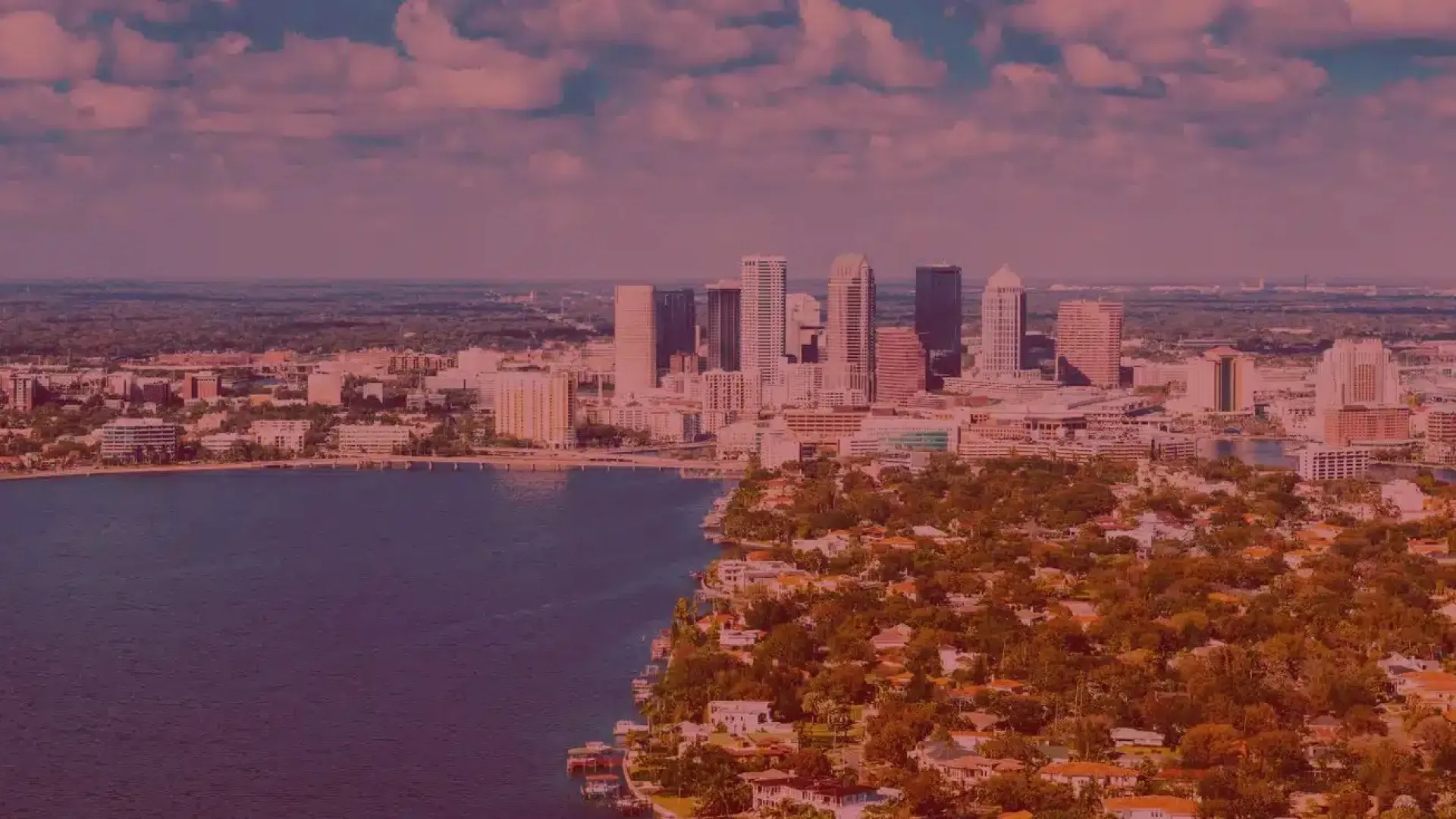 Aerial view of a coastal city skyline with waterfront homes and tall buildings under a partly cloudy sky.