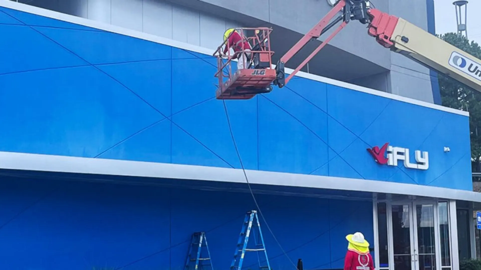 Workers use a boom lift to install signage on the blue exterior wall of an iFLY indoor skydiving center.