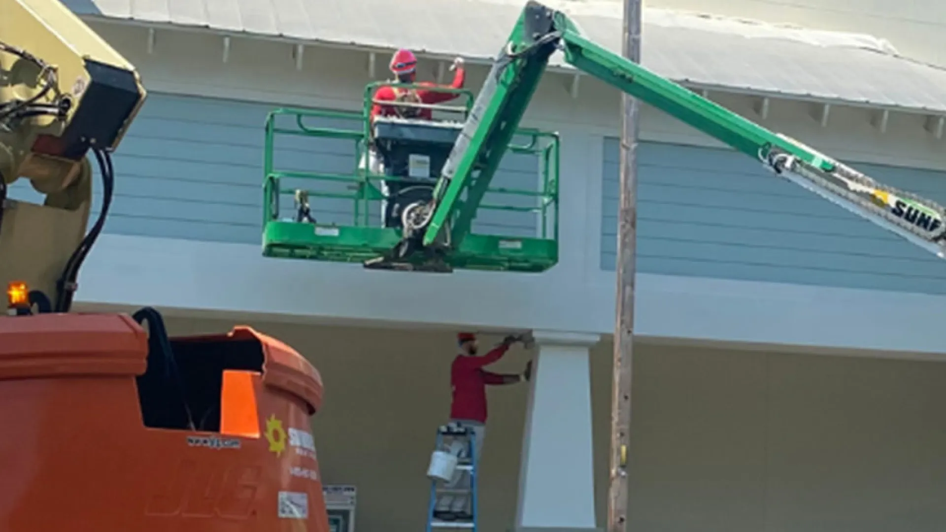 Construction workers operate green boom lift outside light-colored building with palm trees under clear sky