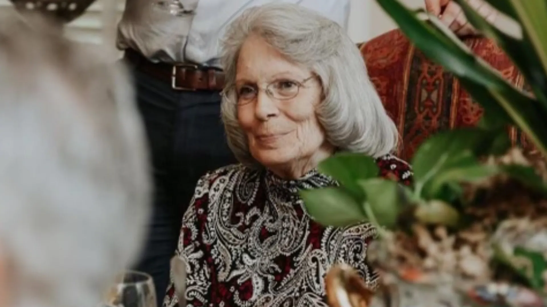 Elderly woman with gray hair and glasses sitting at a table during a social gathering with others.