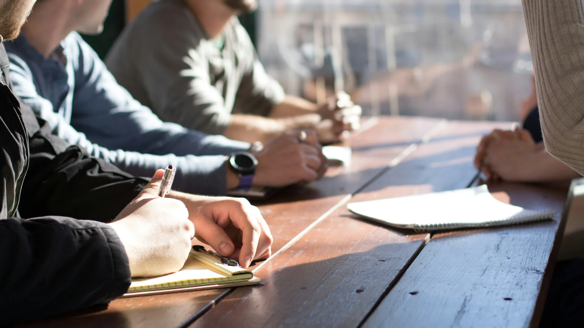 People sitting at a wooden table during a meeting, taking notes on notepads in natural light.