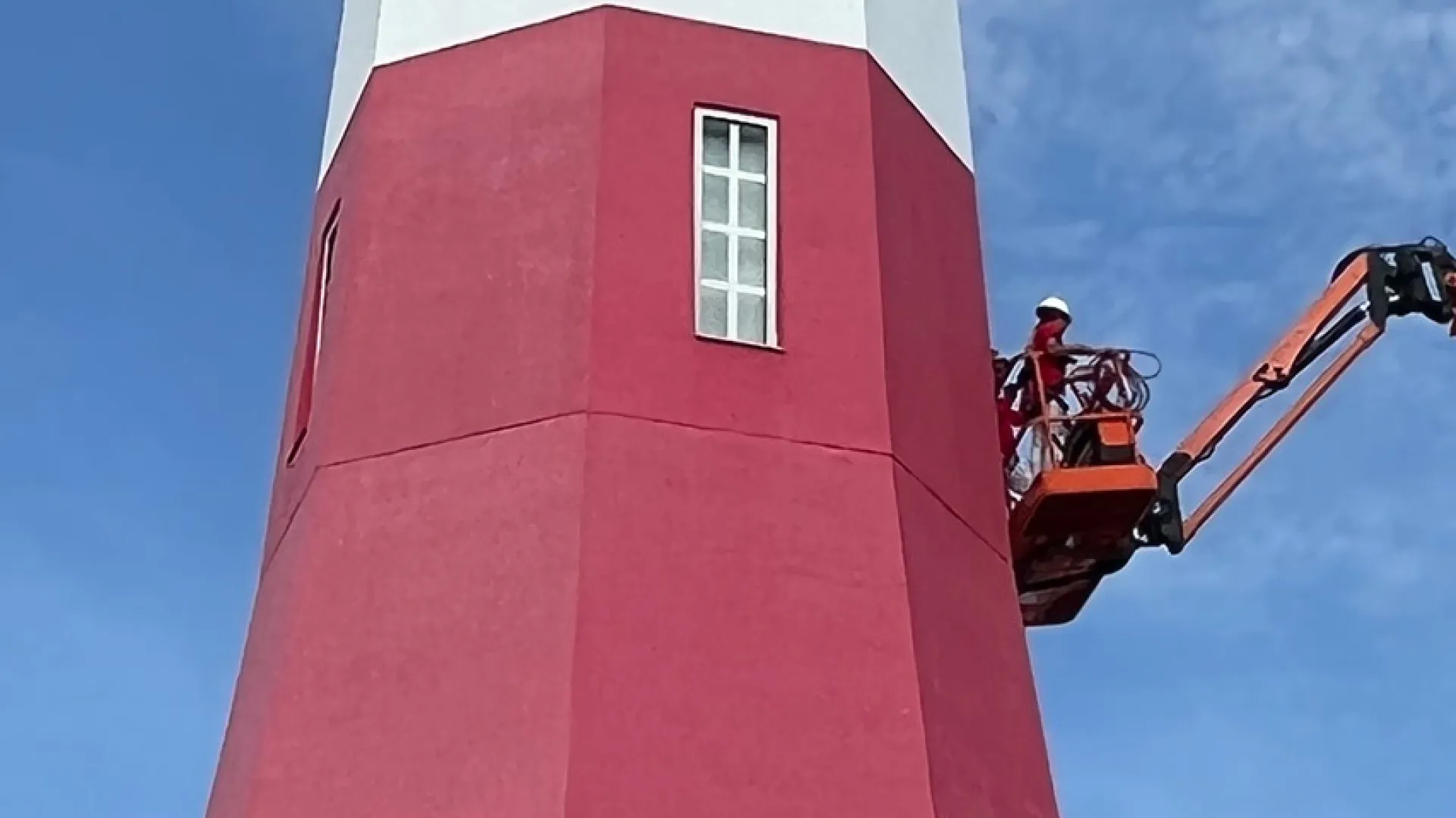 Worker on an orange boom lift painting red and white lighthouse against blue sky background.