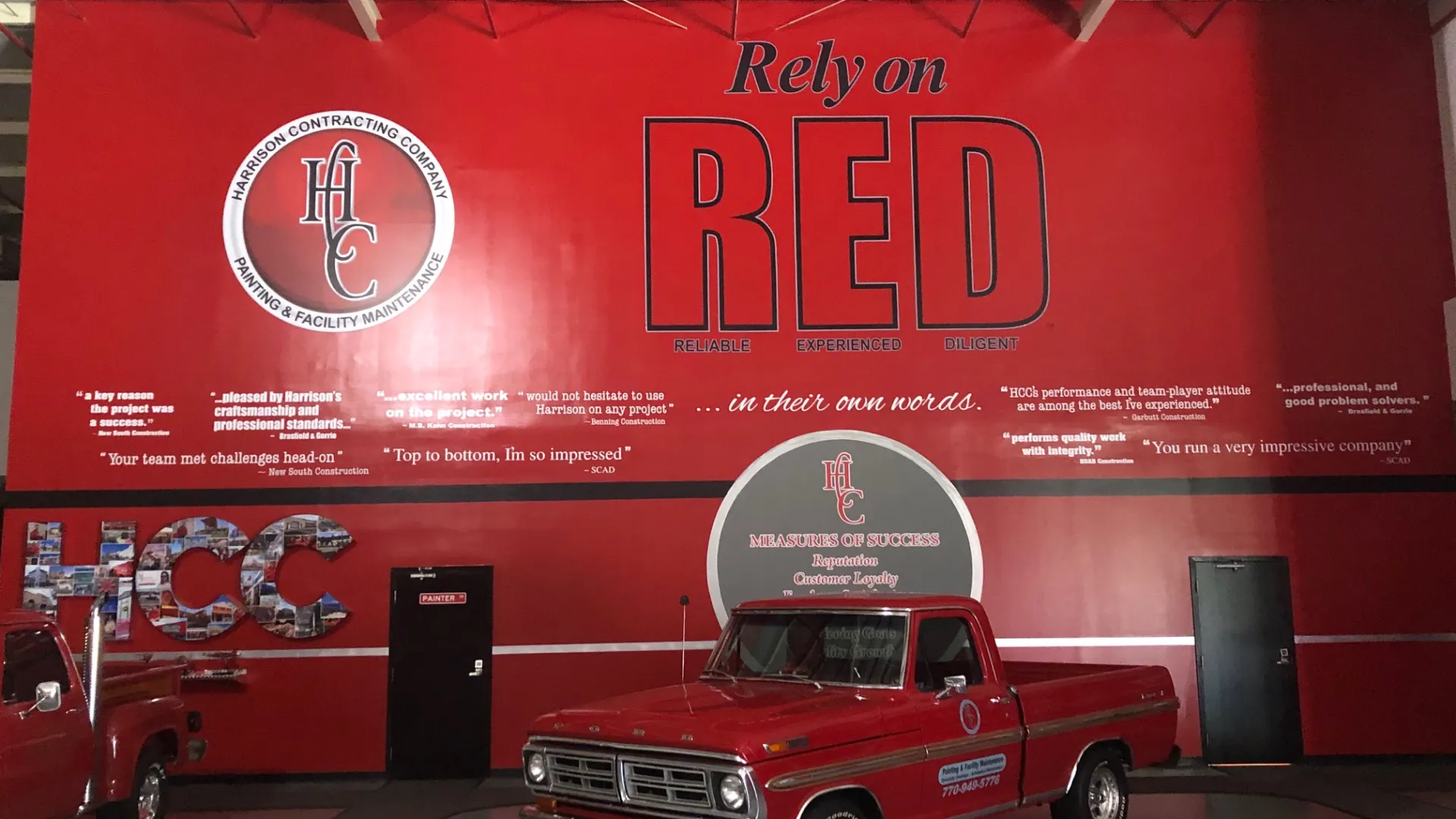 Red wall with 'Rely on RED' slogan, Harrison Contracting logo, and classic red pickup truck in warehouse.