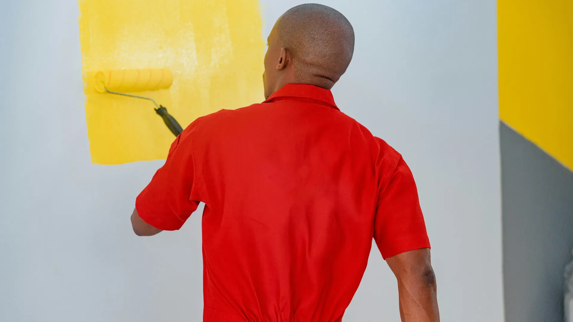 Man in red coveralls painting a white wall with bright yellow paint using a roller brush indoors.