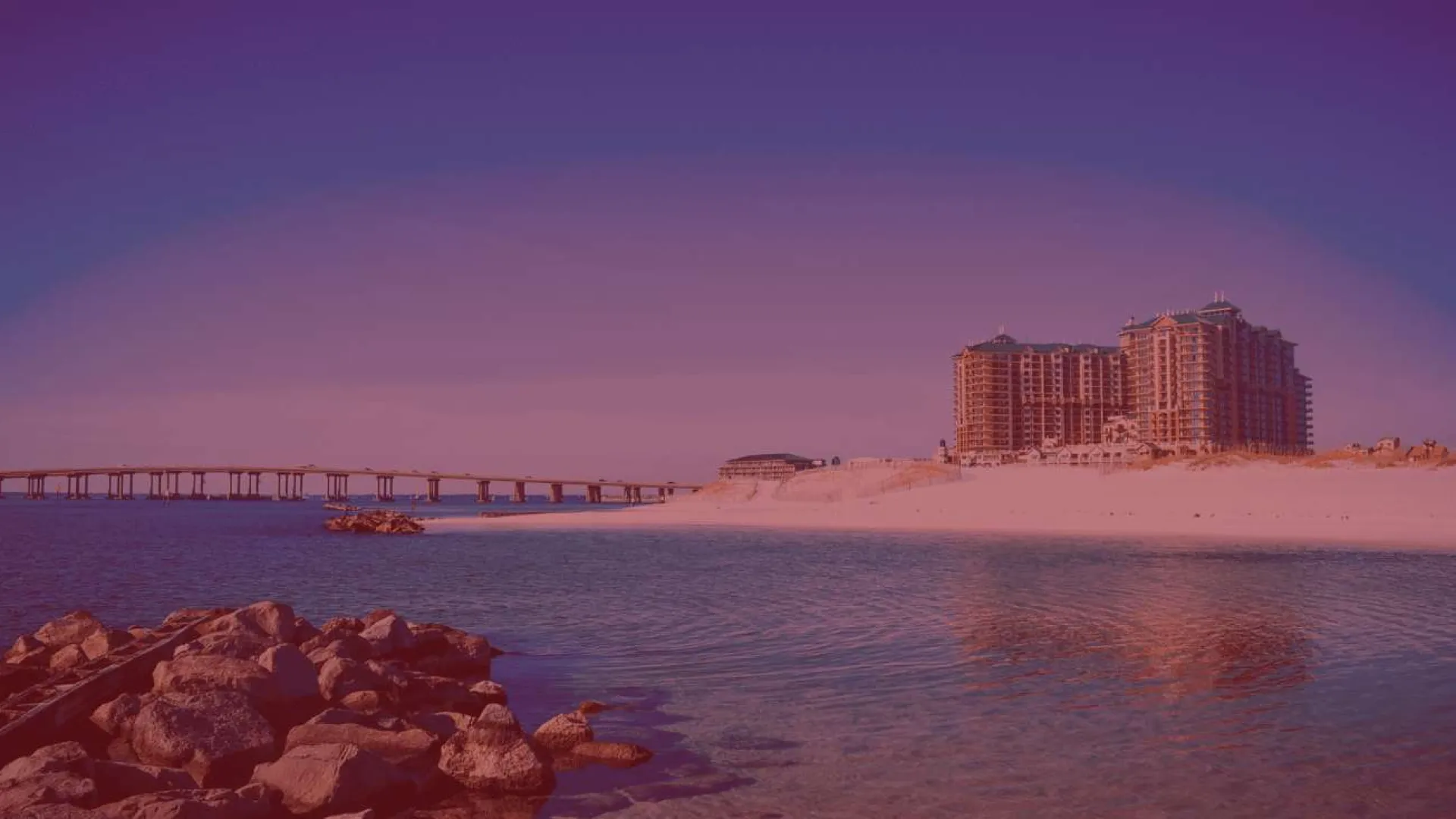Coastal scene with calm water, rocky shoreline, sandy beach, a long bridge, and tall buildings at sunset.