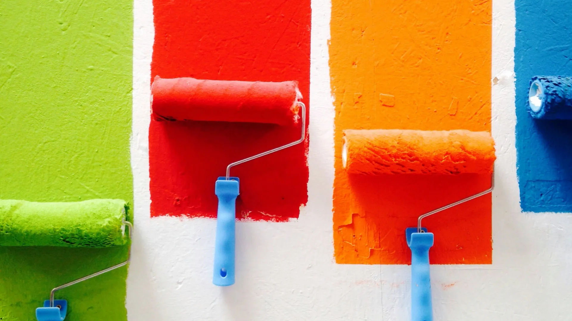 Four paint rollers with blue handles applying vertical stripes of green, red, orange, and blue paint on a white wall.