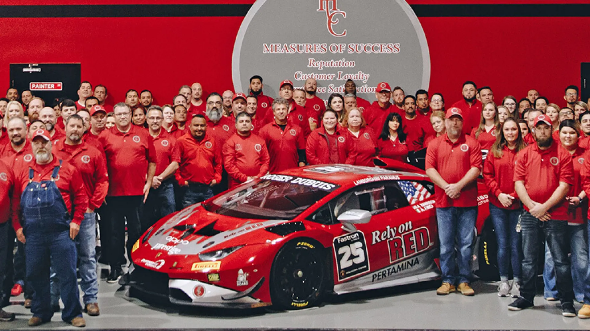 Large team in red uniforms posing with a red racing car inside a showroom featuring HCC logo and signage.