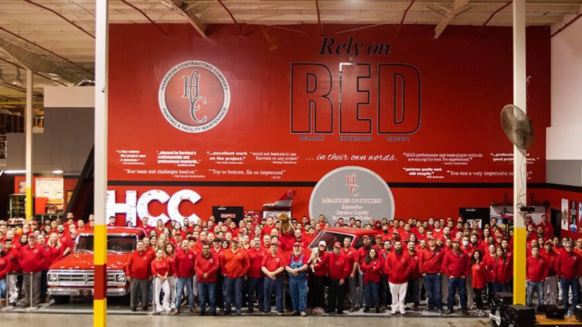 Large group of employees in red shirts posing in front of red wall with HCC industrial contracting slogans and logos