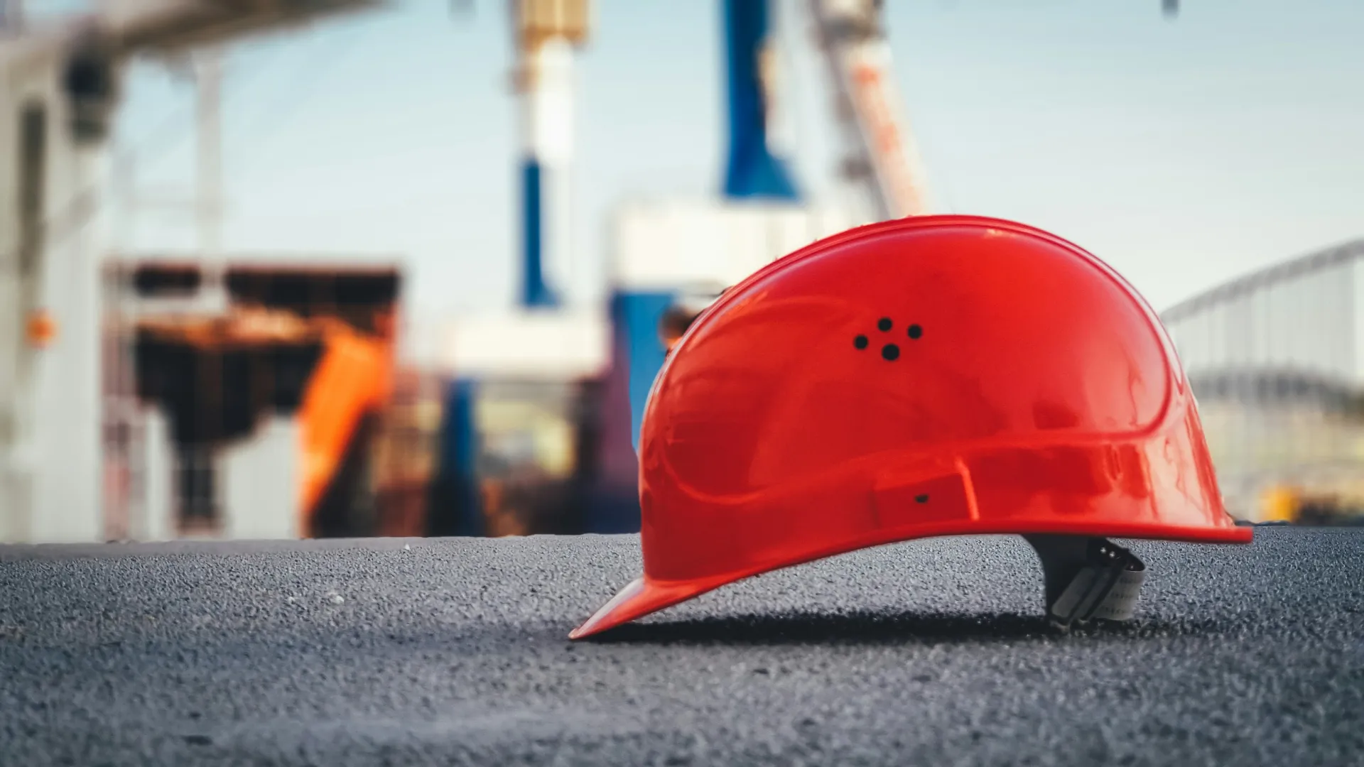 Red construction helmet resting on asphalt at a busy construction site with workers and cranes blurred in background
