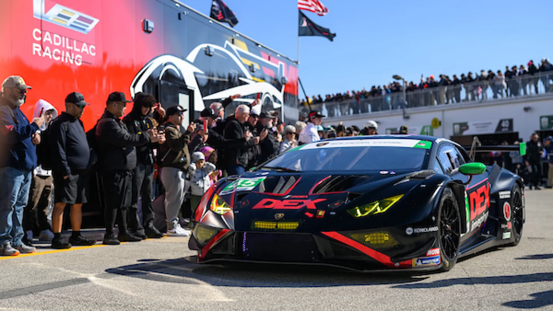 Black and red DEX racing car with yellow headlights driving past a crowd by Cadillac Racing truck.