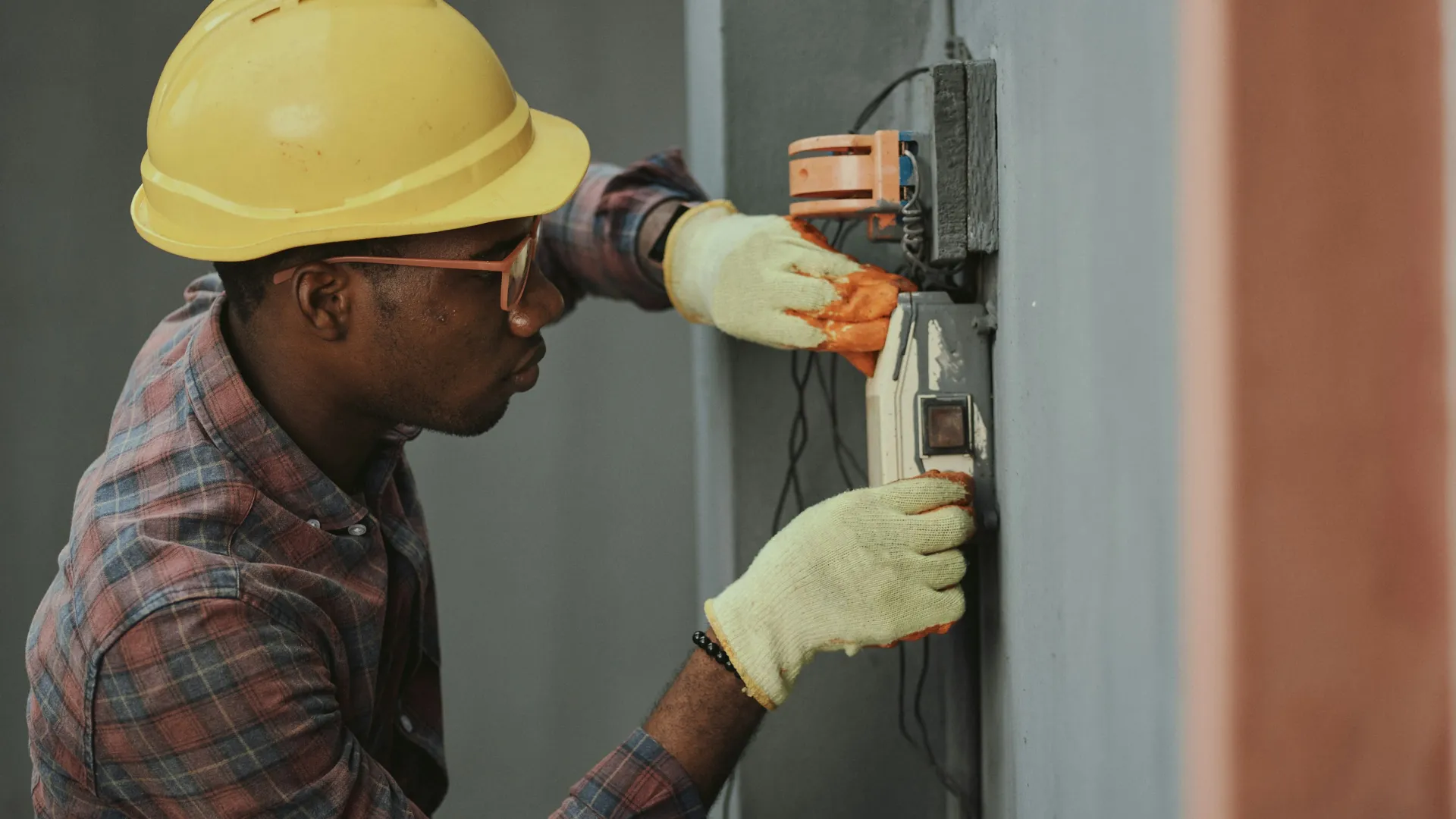 Electrician wearing hard hat and gloves working on electrical panel with tools on grey wall.