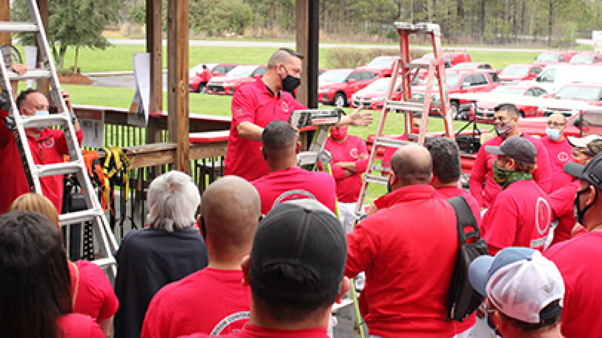 Group of people wearing red shirts and masks gathered outdoors around ladders for a safety briefing or training.
