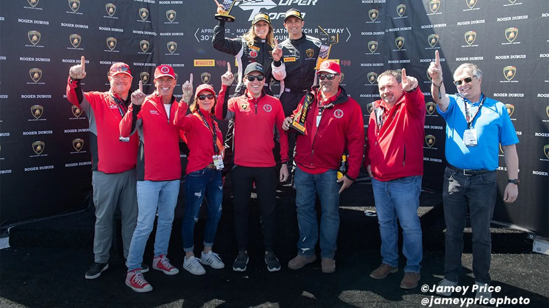 Winning racing team celebrating on podium holding trophy with team members showing number one gesture.