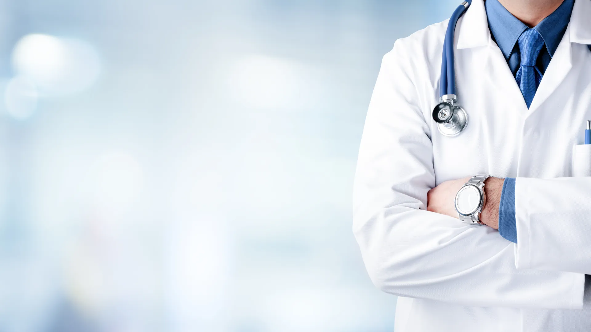Doctor in white coat with stethoscope and arms crossed against blurred medical background