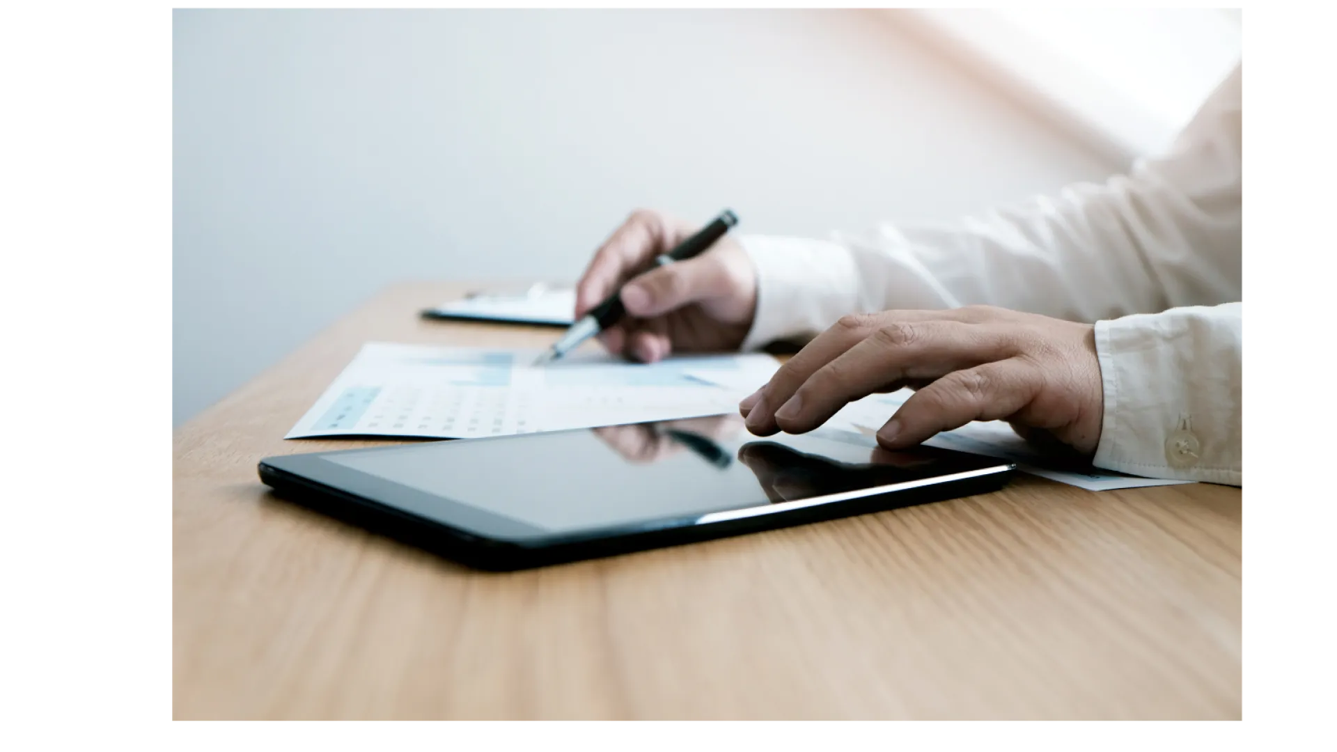 Person in white shirt using tablet and reviewing documents with pen at wooden desk in bright room.