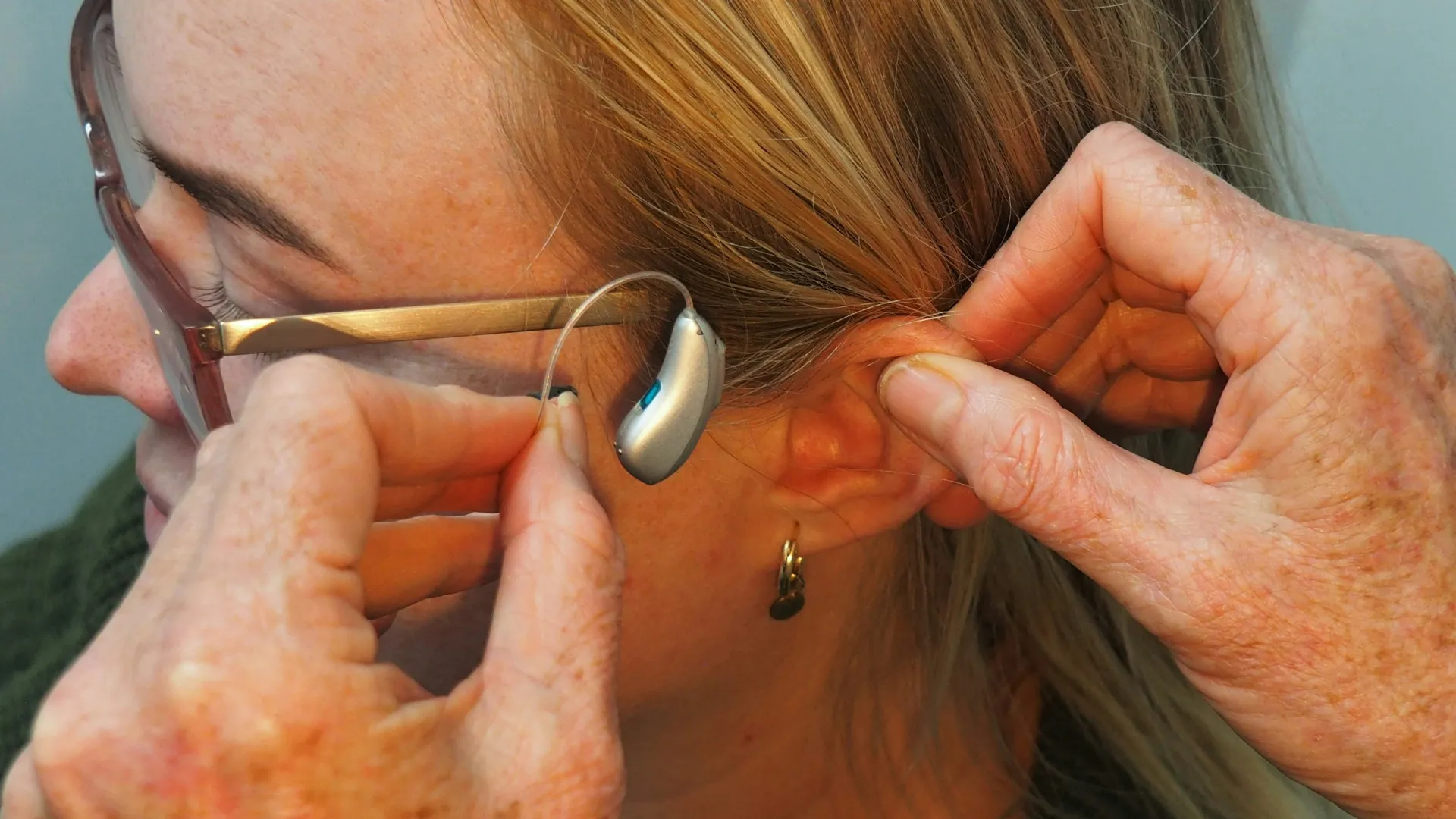 Close-up of a woman fitting a hearing aid behind her ear, adjusting it carefully with both hands.