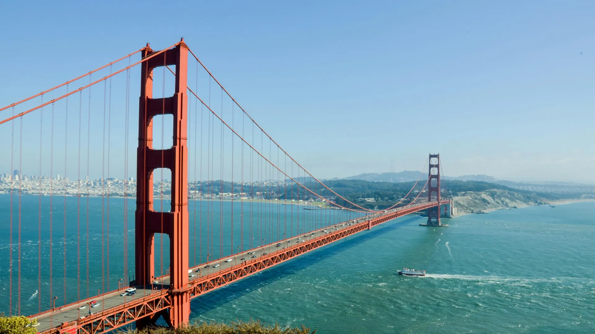 Golden Gate Bridge spanning a bay, surrounded by hills and a clear blue sky, with boats on the water.