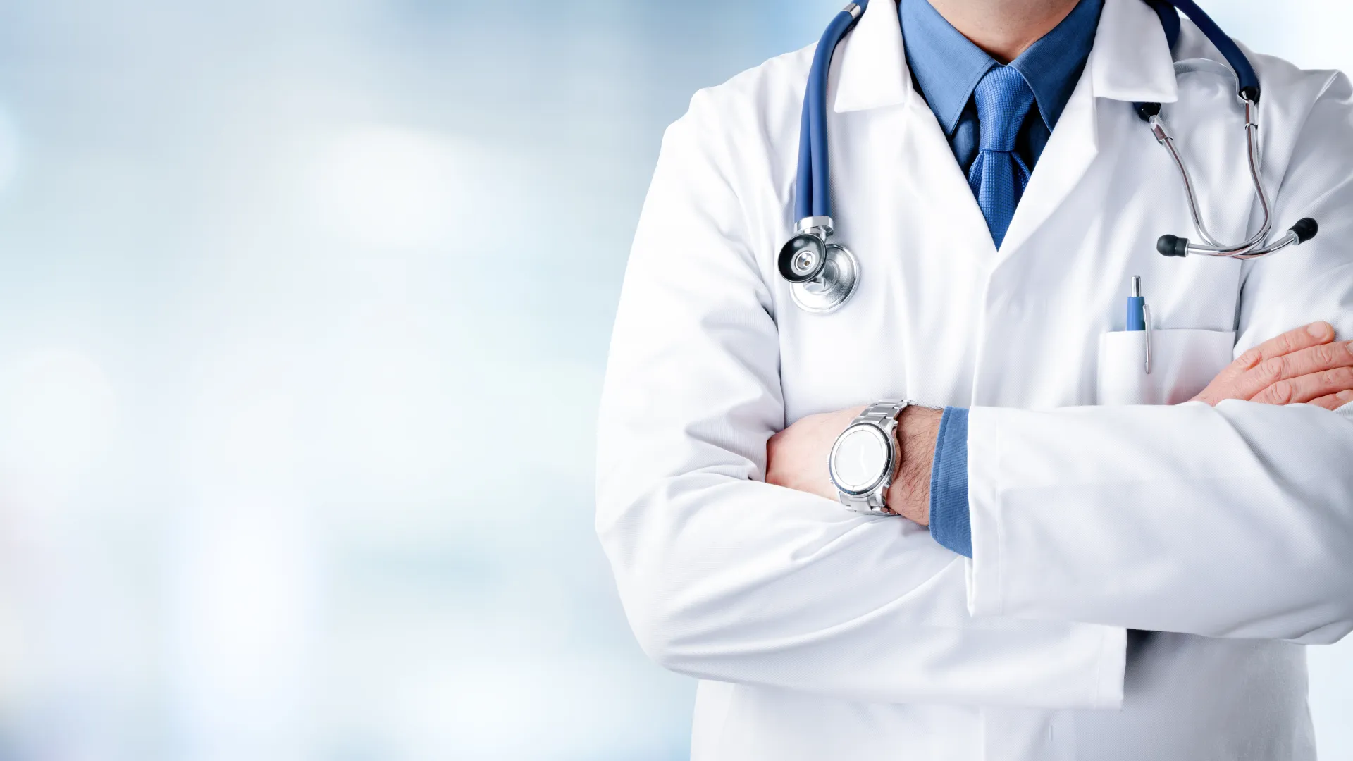 Confident doctor with folded arms wearing white coat and stethoscope in a bright medical setting.